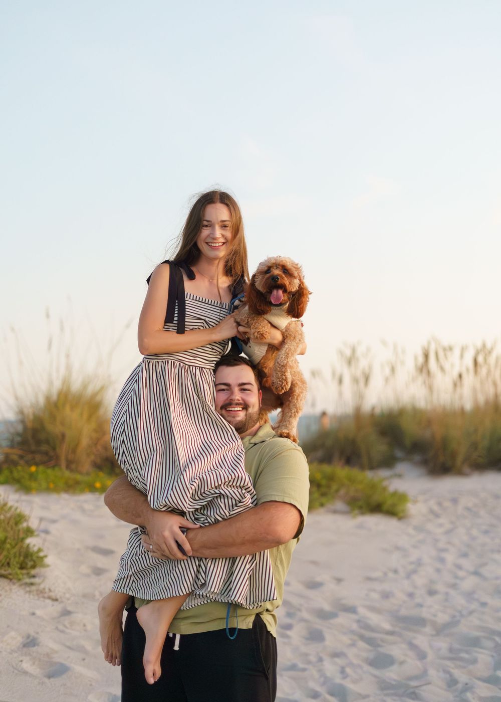 A couple shares playful moments with their small brown dog on a sandy beach at sunset.