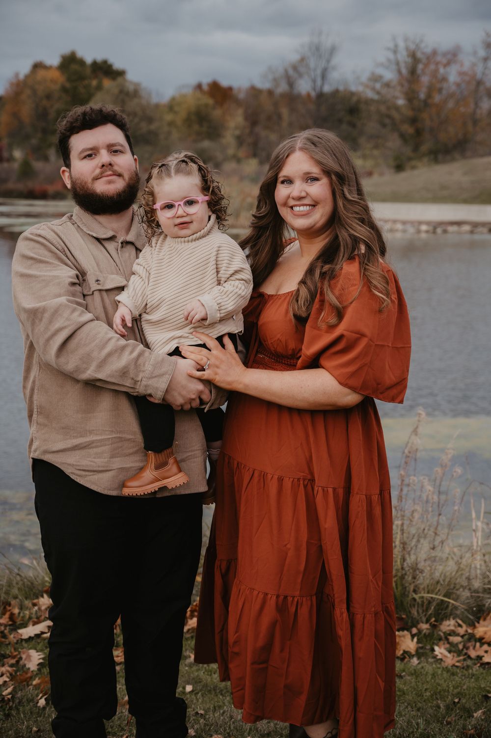 Smiling family of three poses by a lake, dad holding toddler in glasses, mom in rust dress.