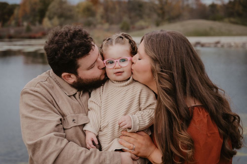 Parents kiss their toddler girl wearing pink glasses on both cheeks outdoors.