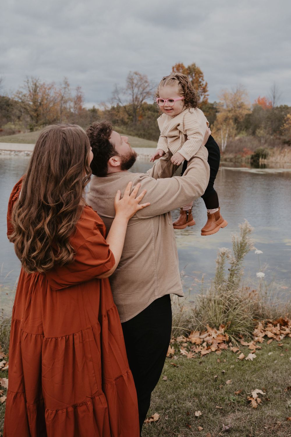 Parents playfully lift their laughing toddler daughter by a lake in autumn.