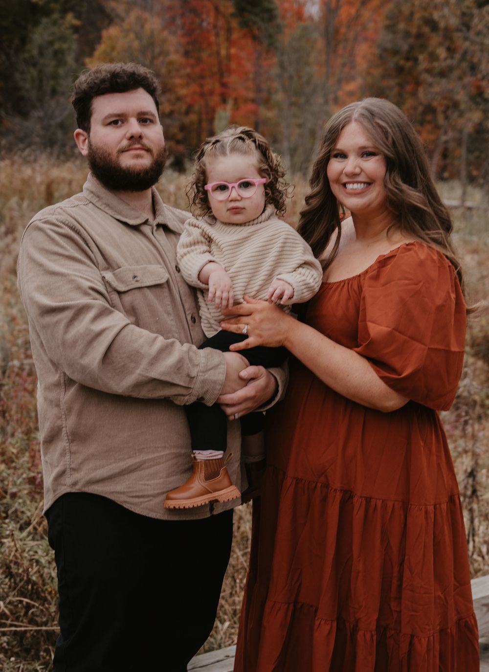 Happy family of three poses in a colorful autumn field, toddler in glasses held by dad.