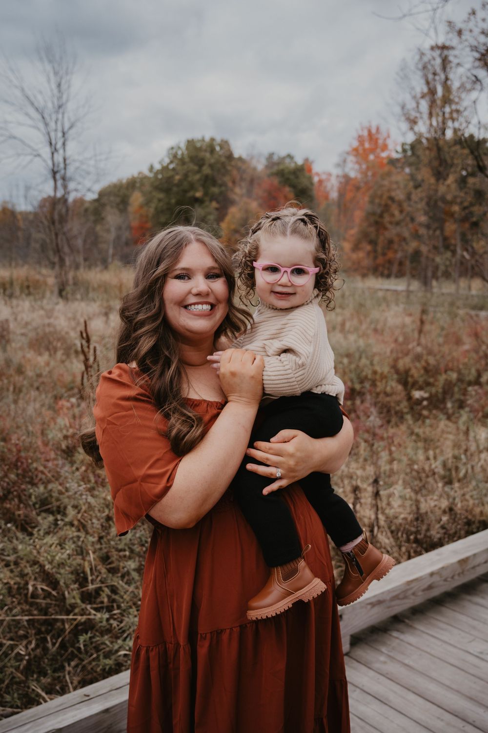 Smiling mom holds toddler girl with pink glasses on a wooden bridge in fall foliage.