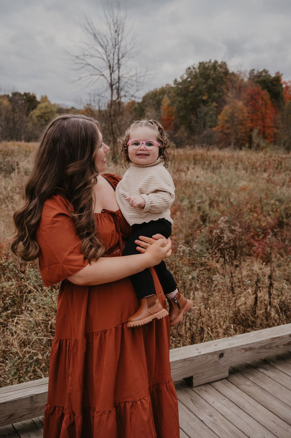 Mom gazes lovingly at her toddler daughter with pink glasses on a fall nature boardwalk.