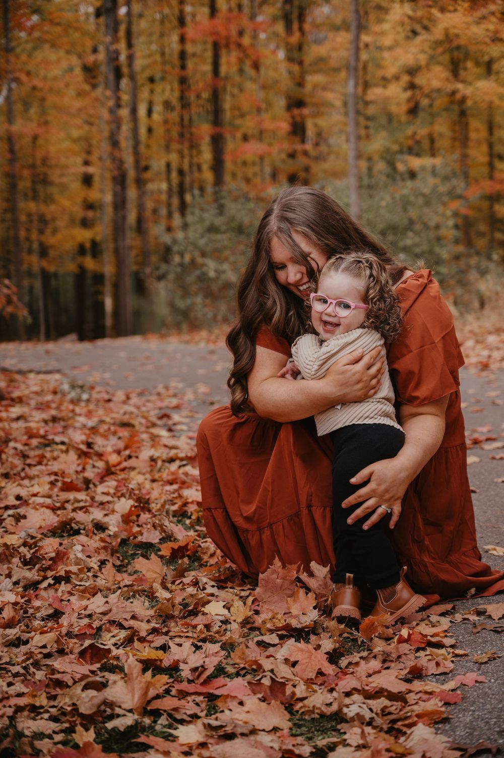 Mother hugging young child in autumn forest with colorful fall foliage and red leaves on ground.