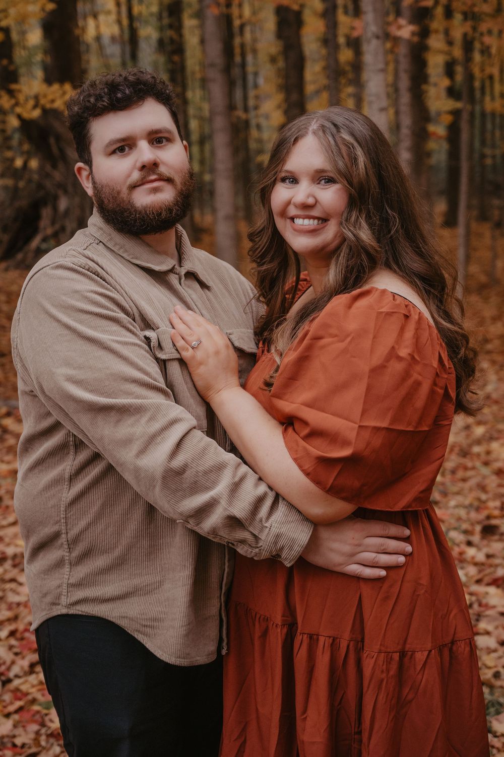 Smiling couple embracing in autumn forest, woman in rust orange dress and man in tan shirt.