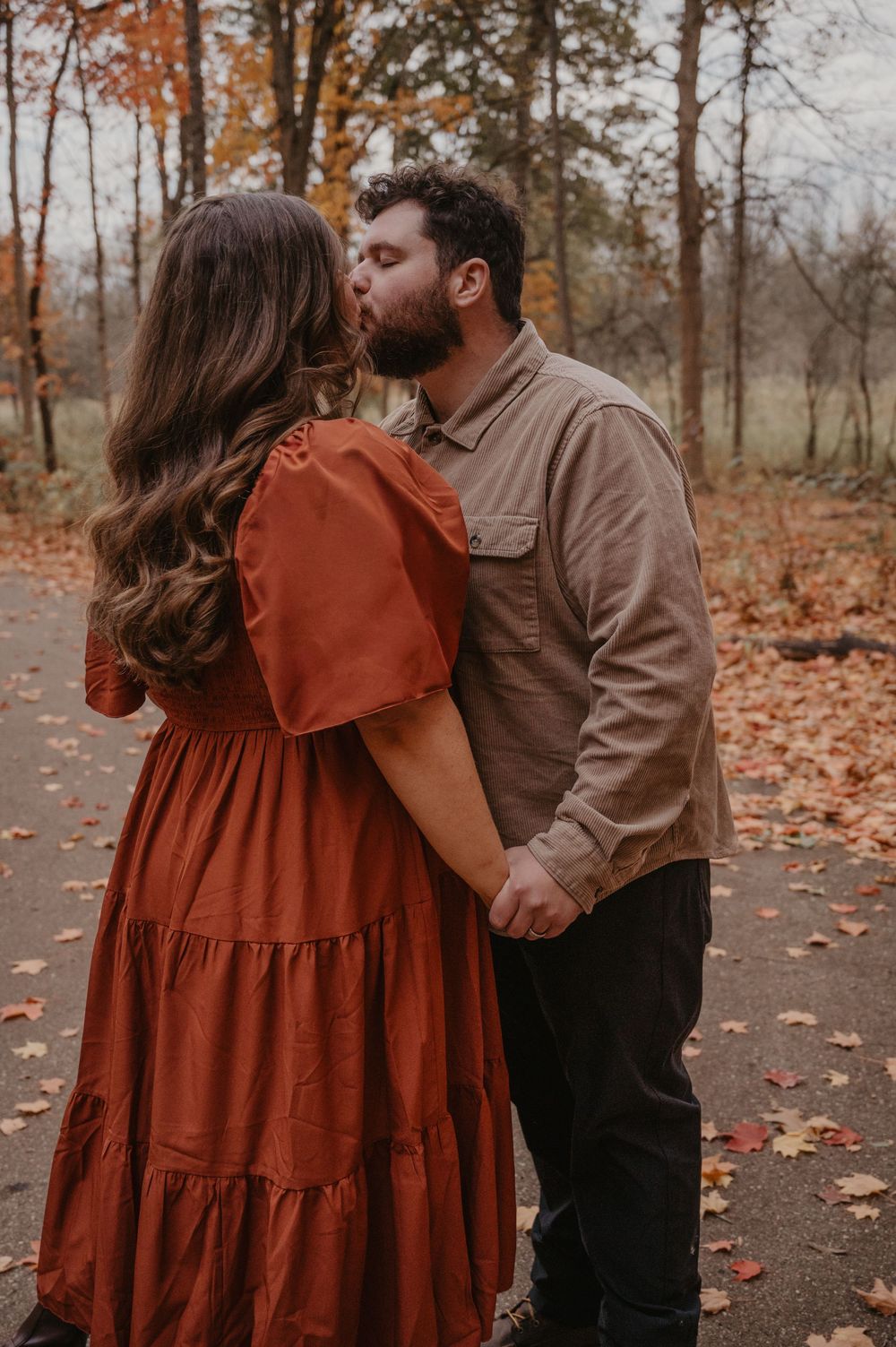 Couple sharing a kiss on a leaf-covered path surrounded by fall foliage.