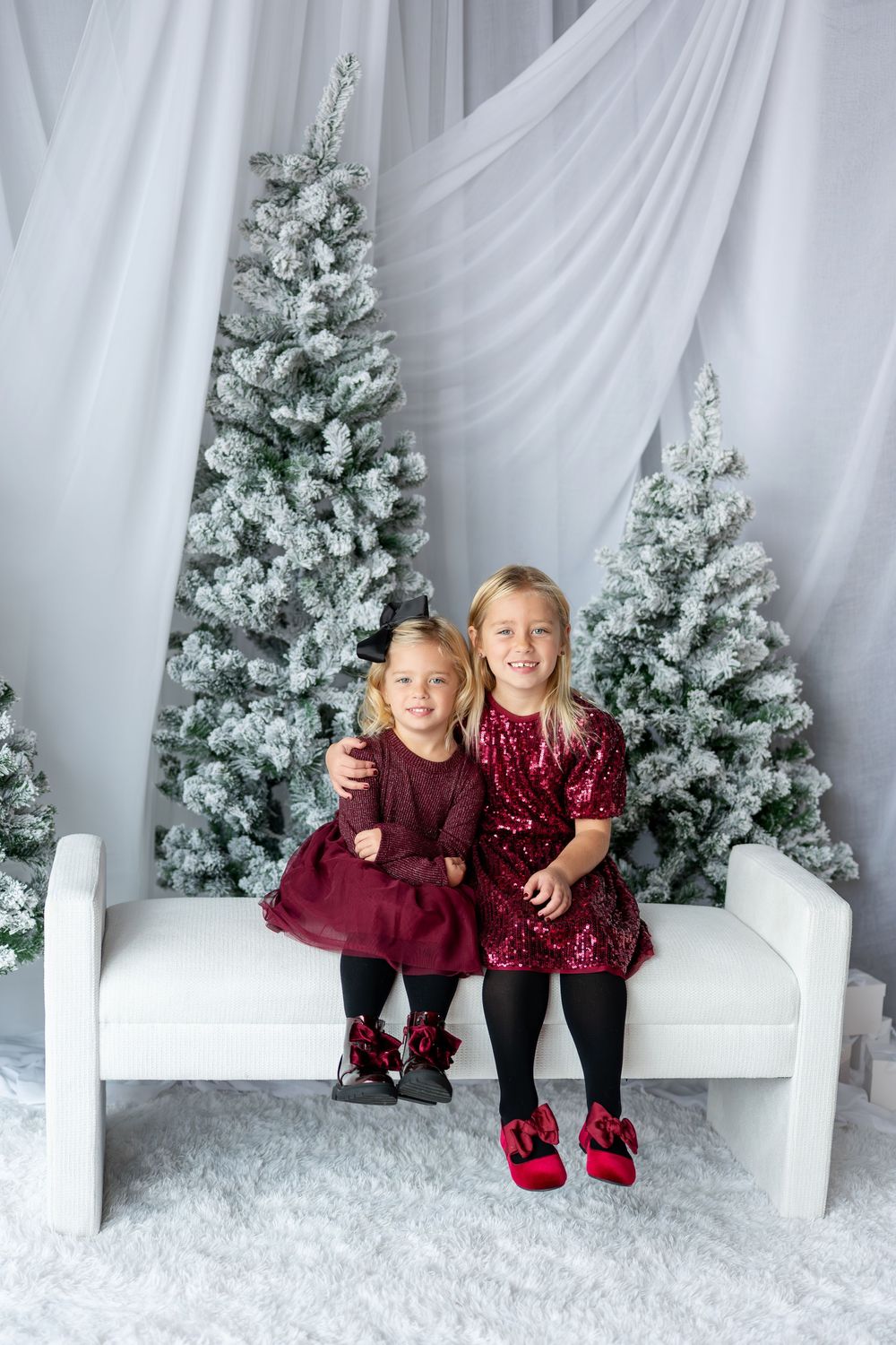 Two young sisters in matching burgundy dresses sitting together on a white bench with snow-flocked Christmas trees.