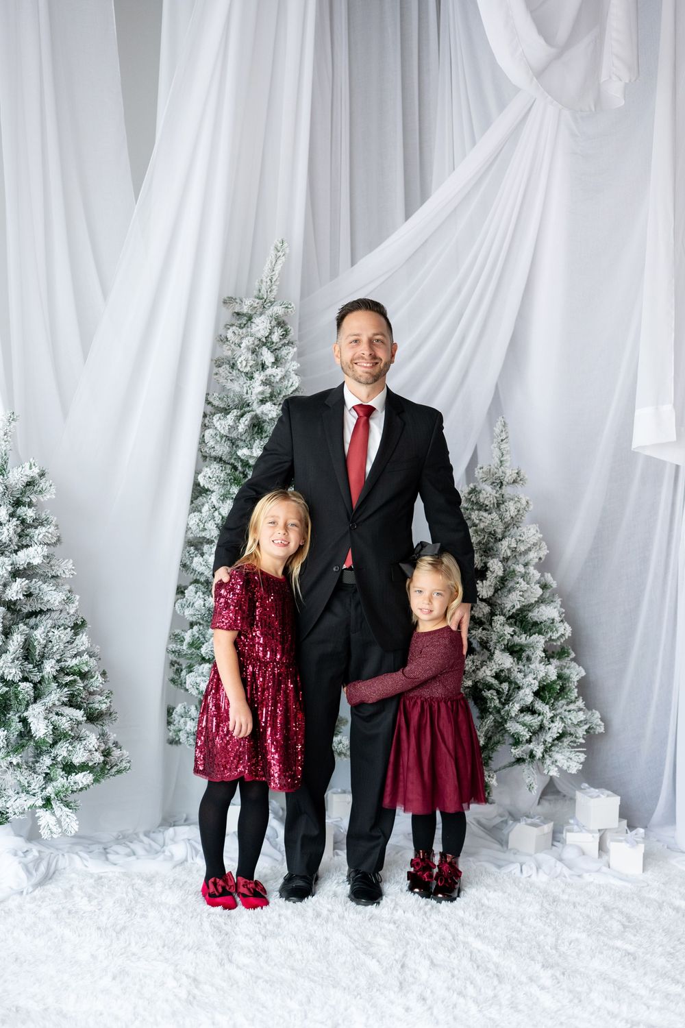 Father in black suit poses with two daughters in burgundy dresses before snowy Christmas trees.