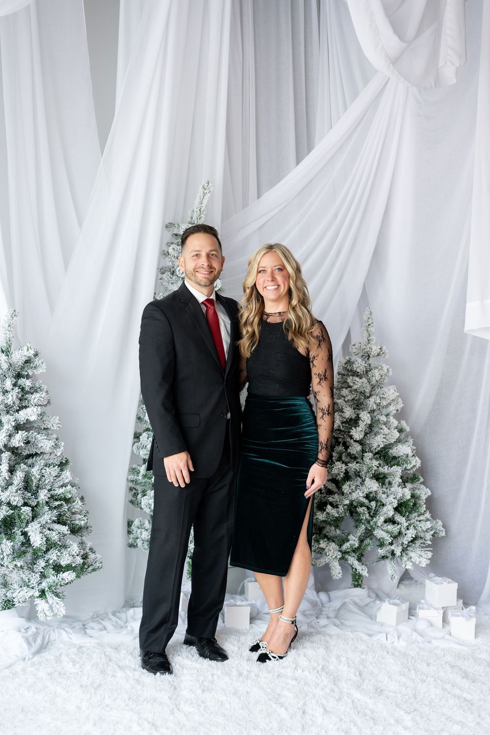 Couple in formal holiday attire poses before snow-flocked Christmas trees with white draped backdrop.