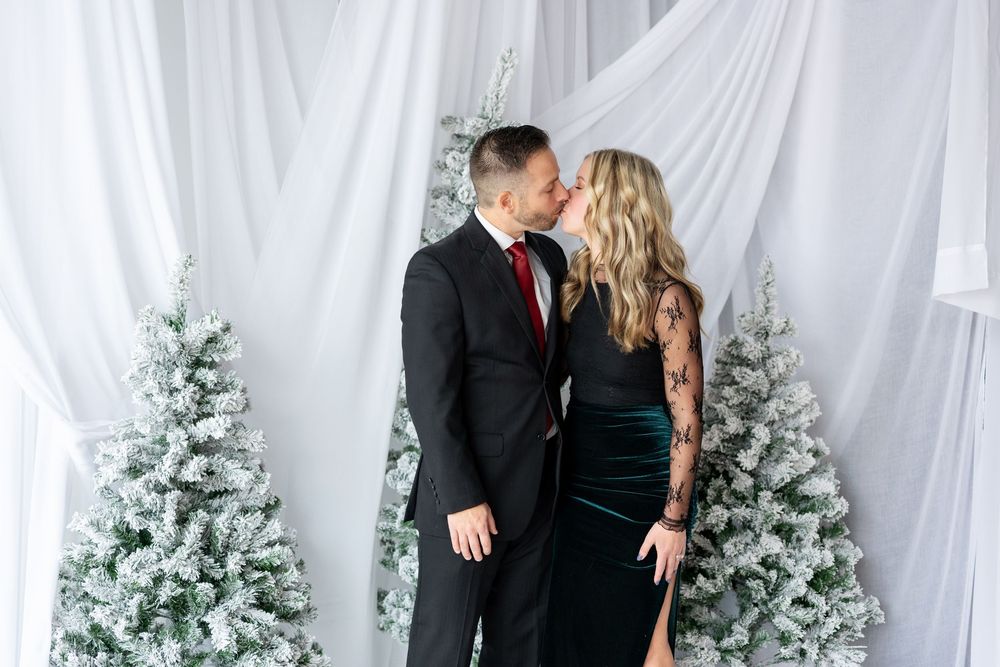 Couple sharing a kiss in formal attire before snow-flocked Christmas trees and white draping.