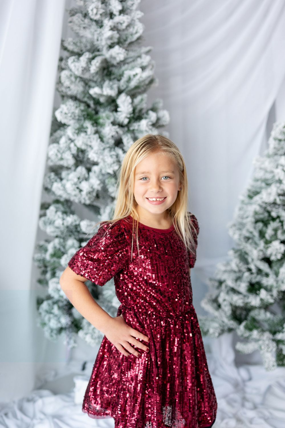 Young blonde girl smiles in a sparkling burgundy sequin dress before snow-flocked Christmas trees.