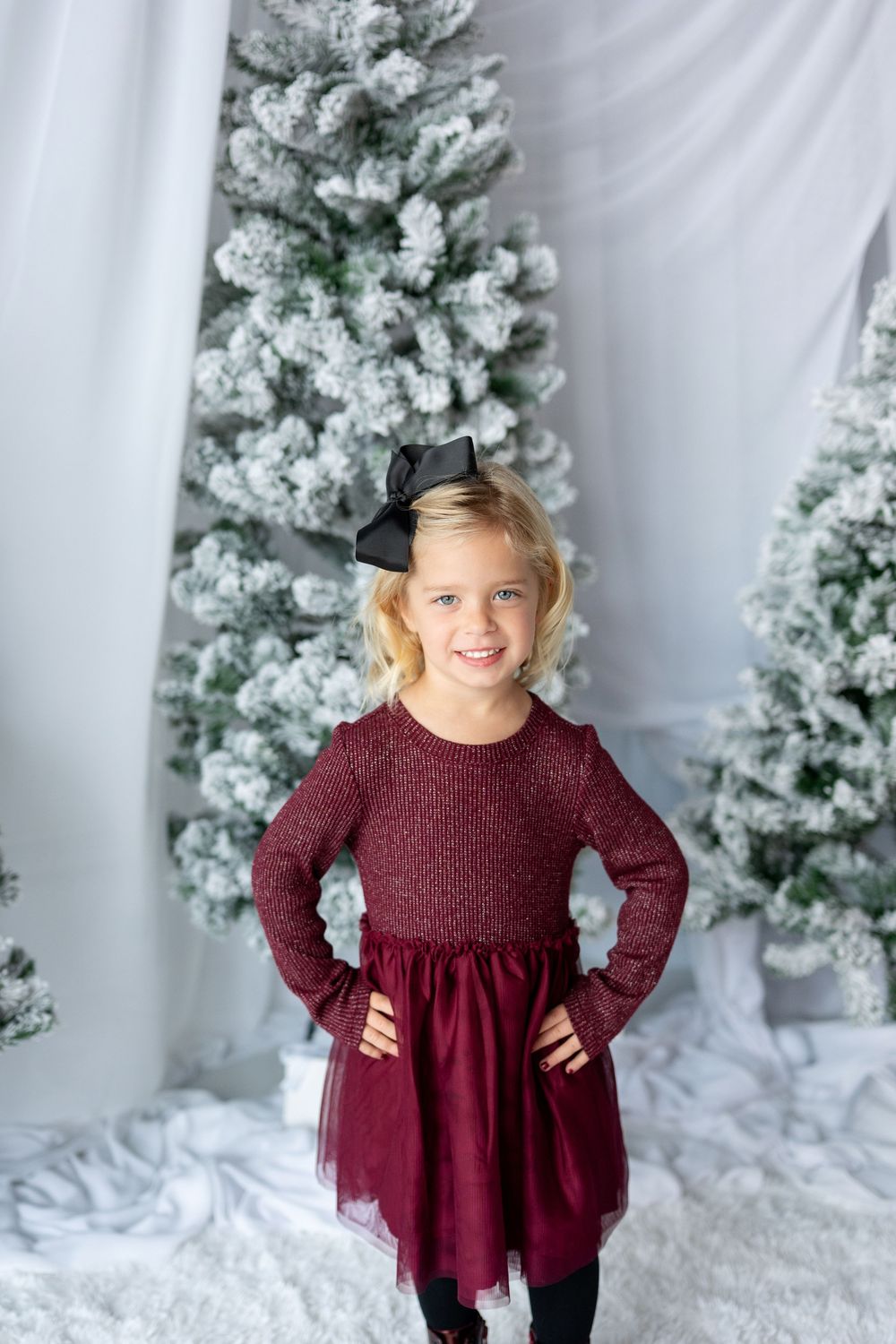 Young blonde girl in burgundy holiday dress with black bow poses before snow-flocked Christmas trees.