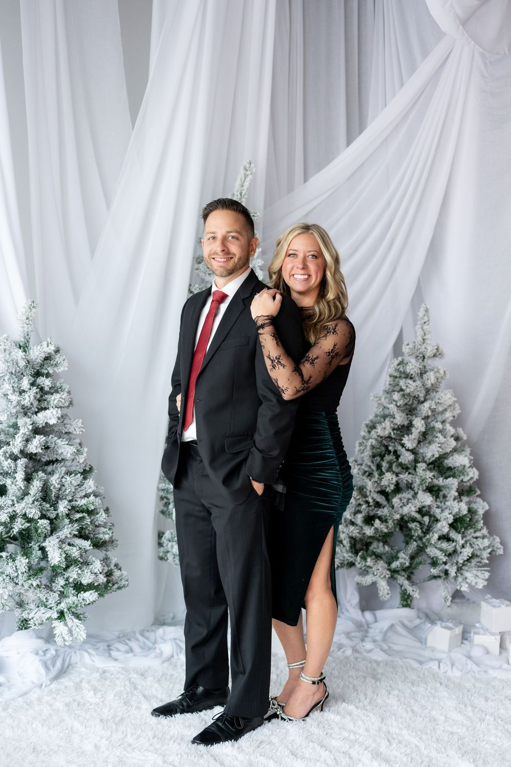 Smiling couple in elegant holiday attire poses before snow-flocked Christmas trees and white draping.