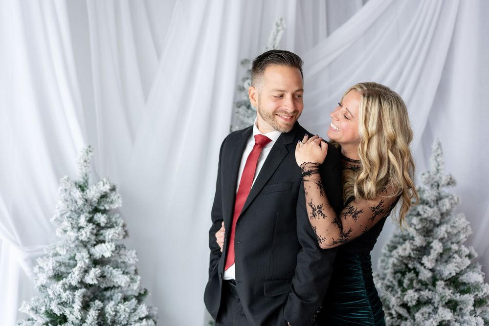 Couple in formal holiday attire laughs together before snow-flocked Christmas trees and white draping.