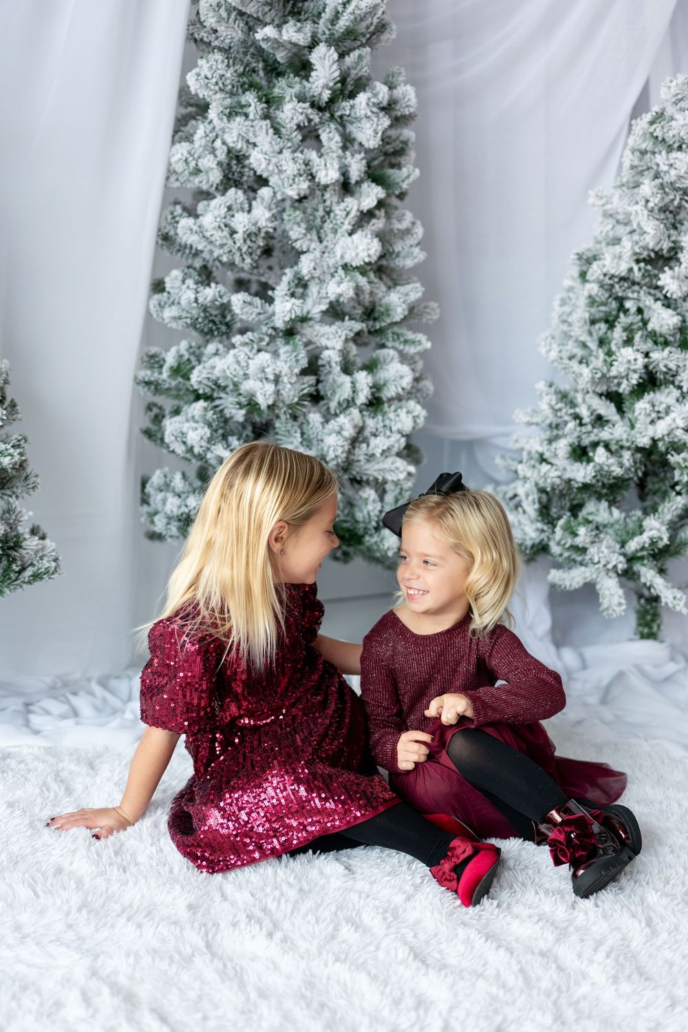 Two young sisters in burgundy holiday dresses sit together chatting before snow-flocked Christmas trees.