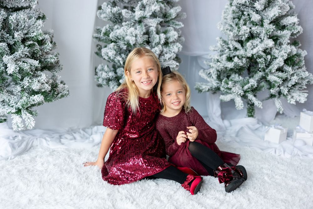 Two smiling sisters in burgundy holiday dresses sit together before snow-flocked Christmas trees.