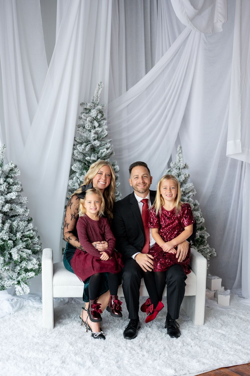 Family of four in holiday attire sitting on a white bench surrounded by snow-flocked Christmas trees for a portrait.