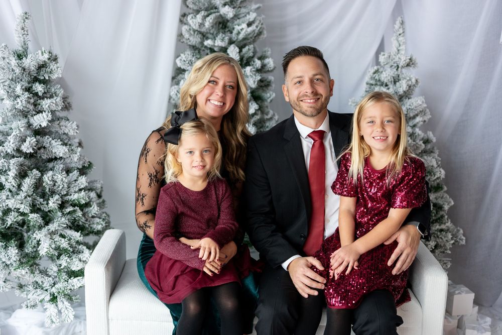 Family of four smiling together on a white bench surrounded by snow-flocked Christmas trees in elegant holiday outfits.