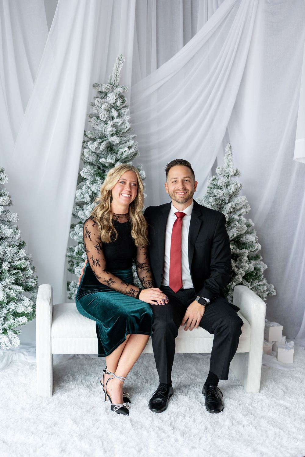 Couple in elegant holiday attire sitting together on a white bench with snow-flocked Christmas trees behind them.