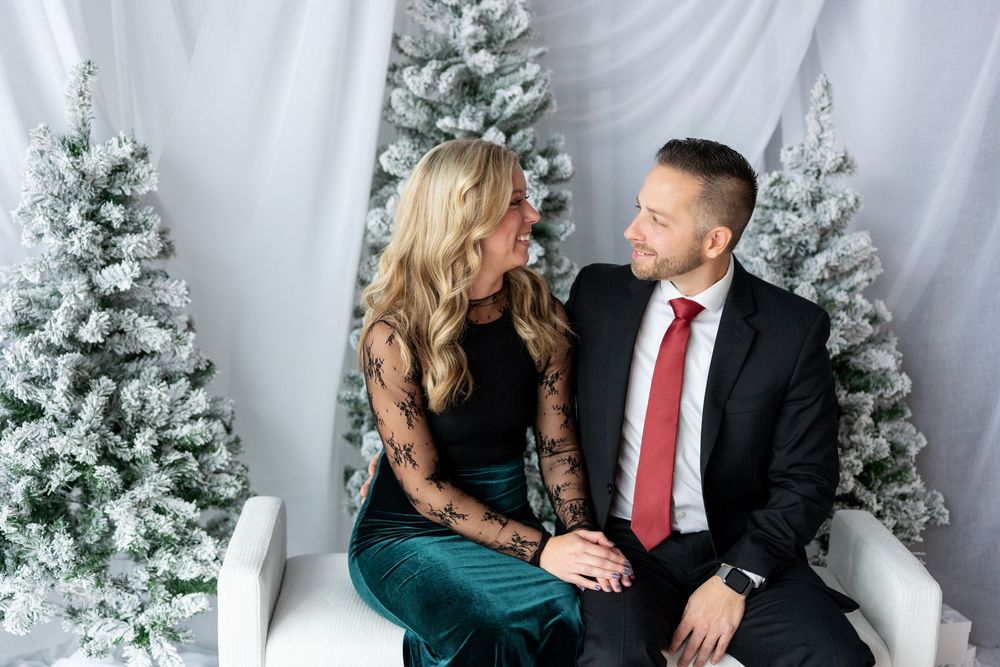 Couple sharing a romantic glance while seated on a white bench with snow-flocked Christmas trees in the background.