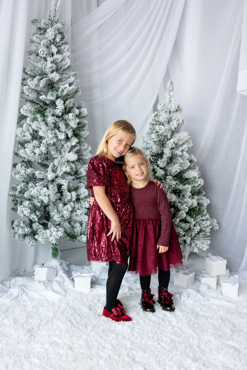 Two young sisters in burgundy dresses hugging and smiling together with snow-flocked Christmas trees and gifts behind them.