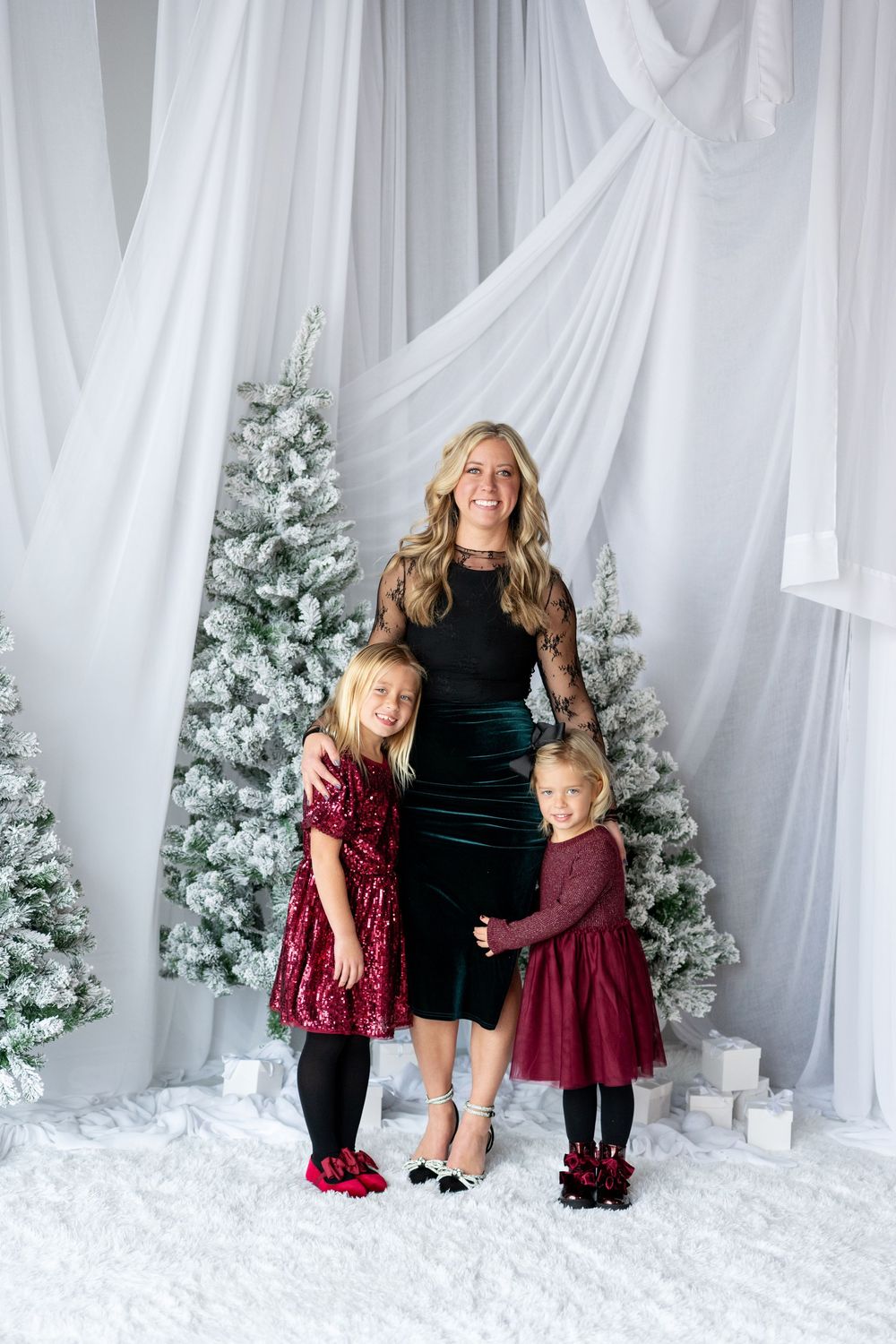 Mother in black velvet dress posing with two young daughters in burgundy dresses before snow-flocked Christmas trees.