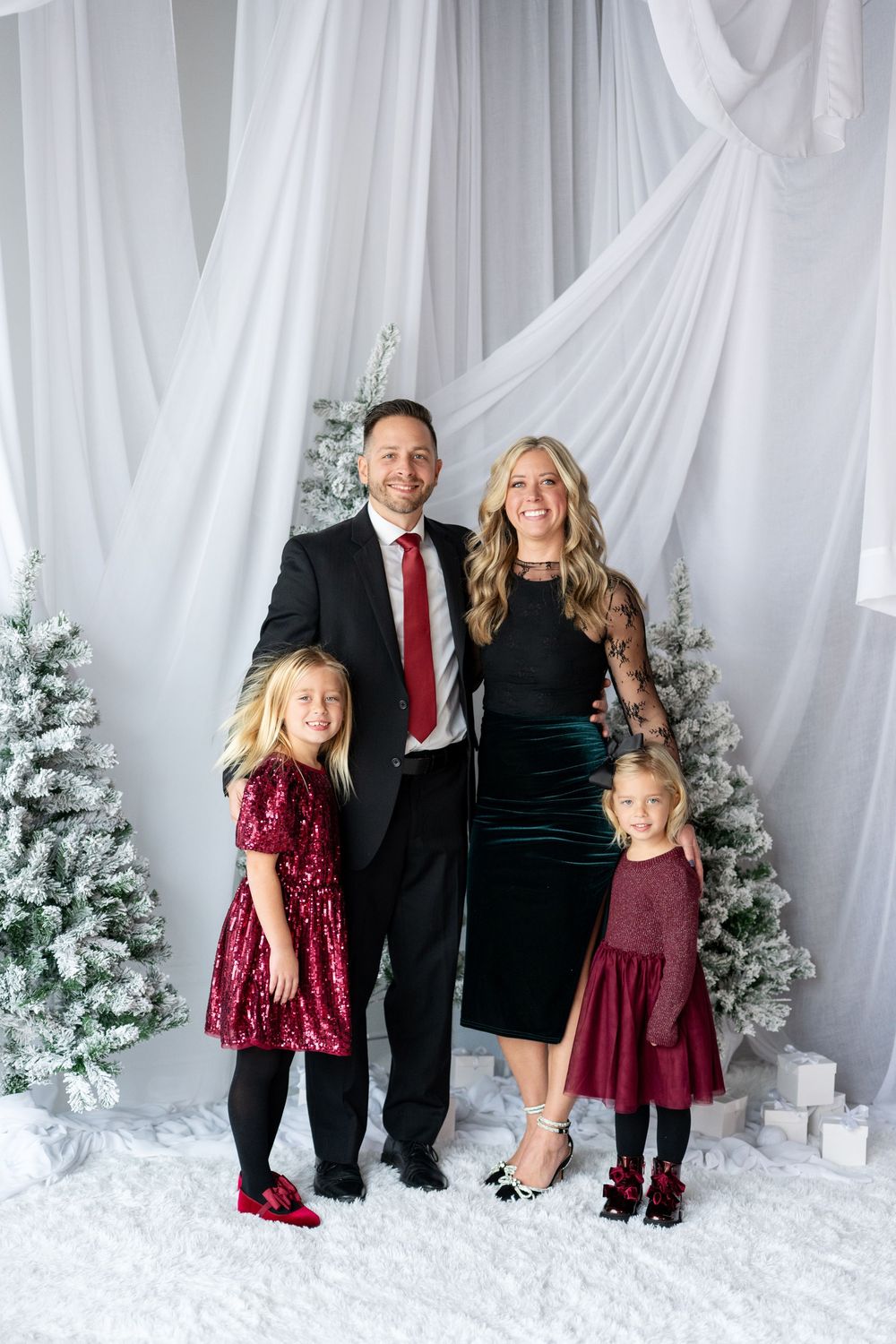 Family of four poses in elegant holiday attire before snow-flocked Christmas trees and white draping.
