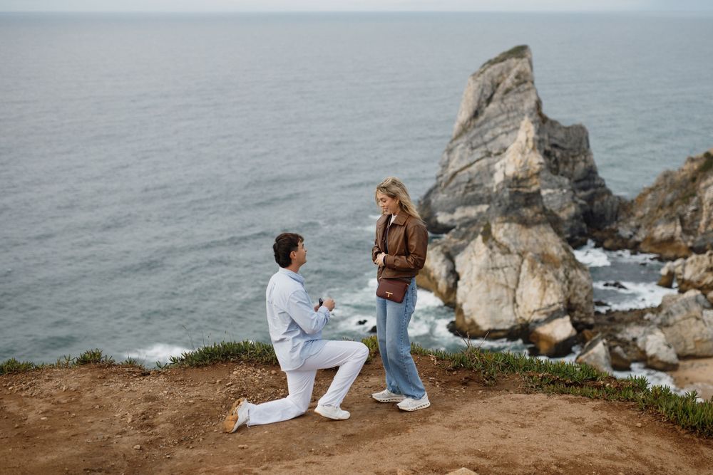 People in white outfits sit along a rocky coastal cliff overlooking the ocean on a cloudy day.