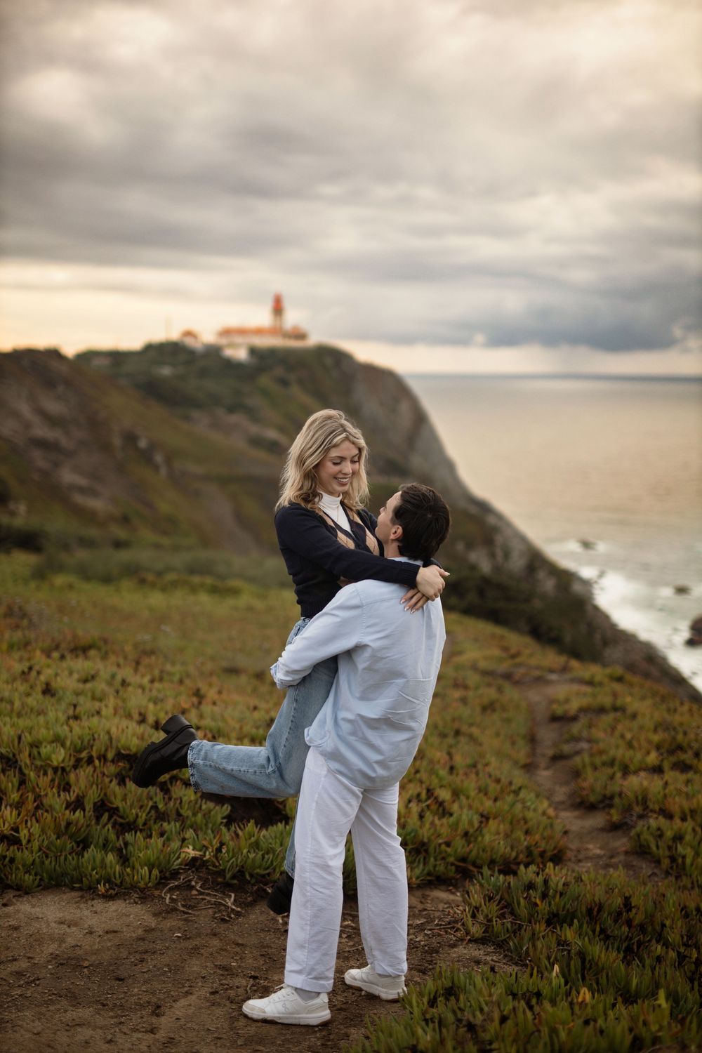 A couple embraces on a scenic coastal cliff at sunset with a lighthouse visible in the background.