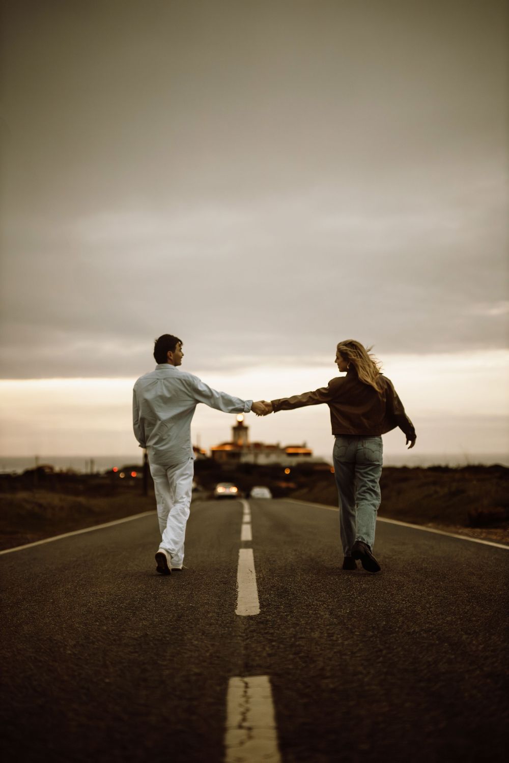 Two people in white clothing stand on opposite sides of an empty desert road under dramatic stormy skies.
