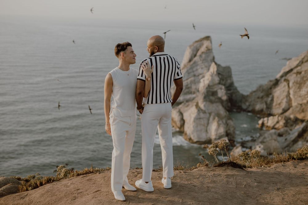 Couple walking along rocky coastal cliffs at sunset with ocean views in the background.