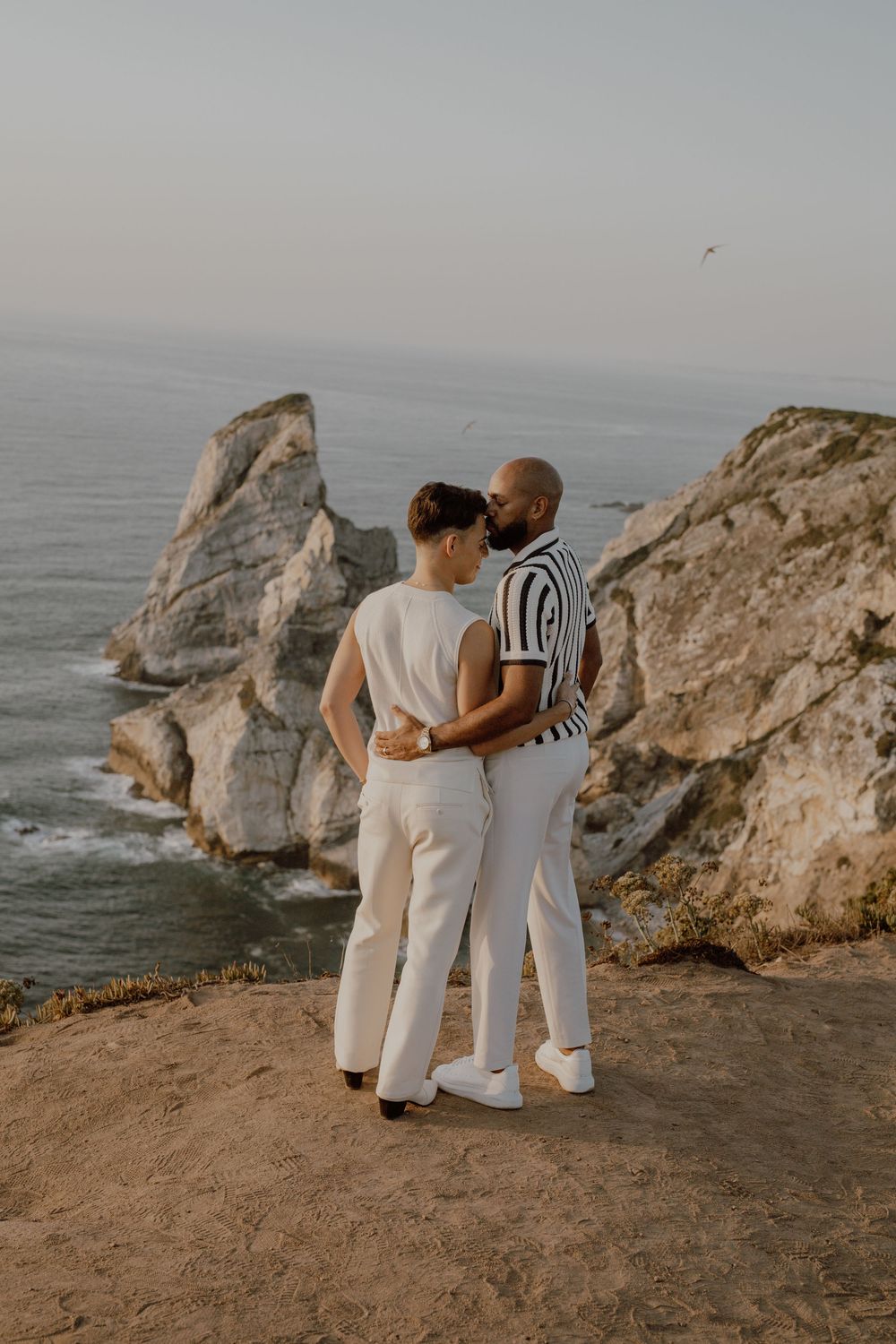 Three people in light-colored outfits embrace while standing on coastal cliffs overlooking the ocean at sunset.