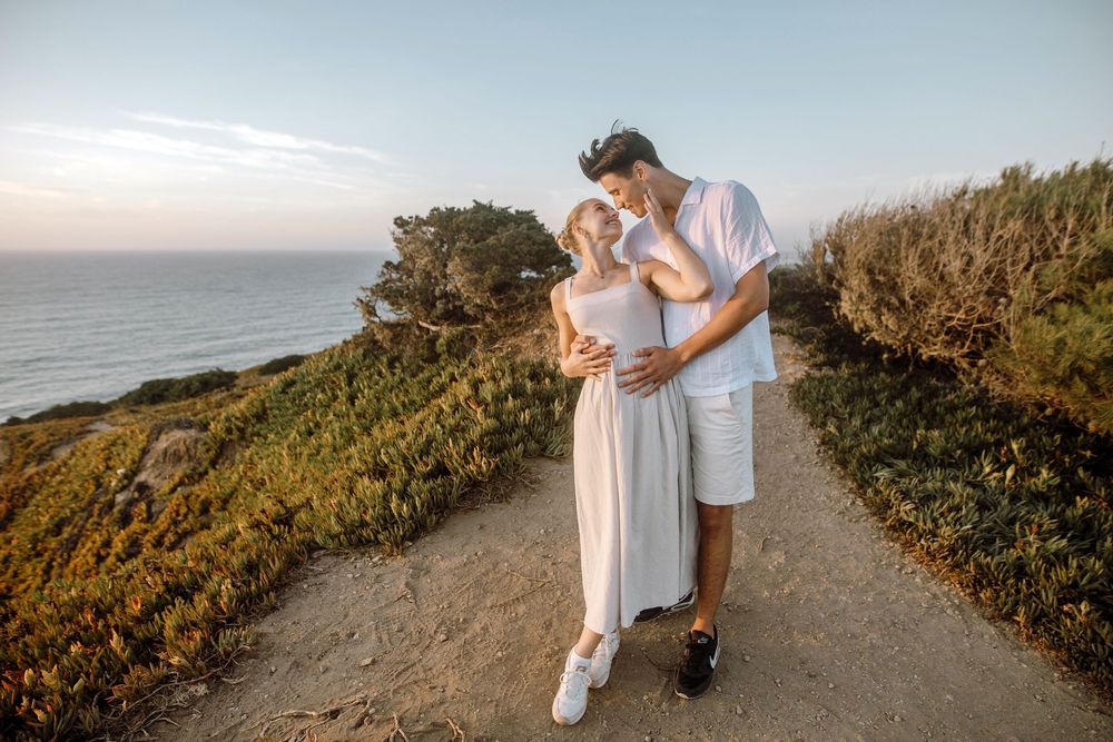 A couple embraces on a scenic coastal path during golden hour overlooking the ocean and cliffside vegetation.