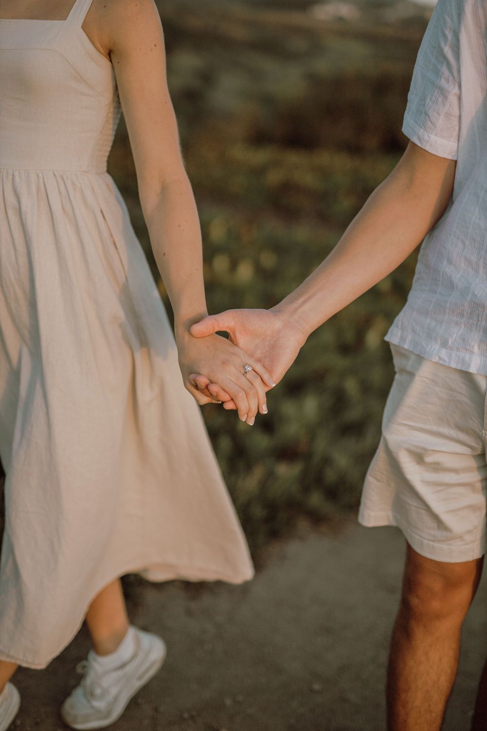 A couple holding hands wearing light summer clothing and sneakers against a natural outdoor backdrop.