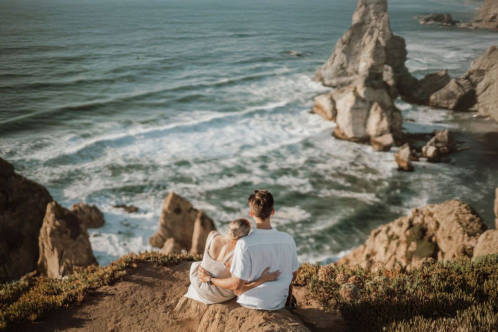 Couple enjoying a romantic sunset moment on coastal cliffs overlooking the ocean waves below.