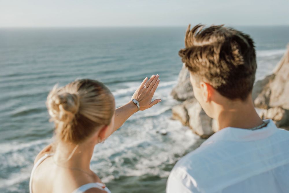 Two people wave while standing at the edge of a scenic coastal cliff with crashing waves below.