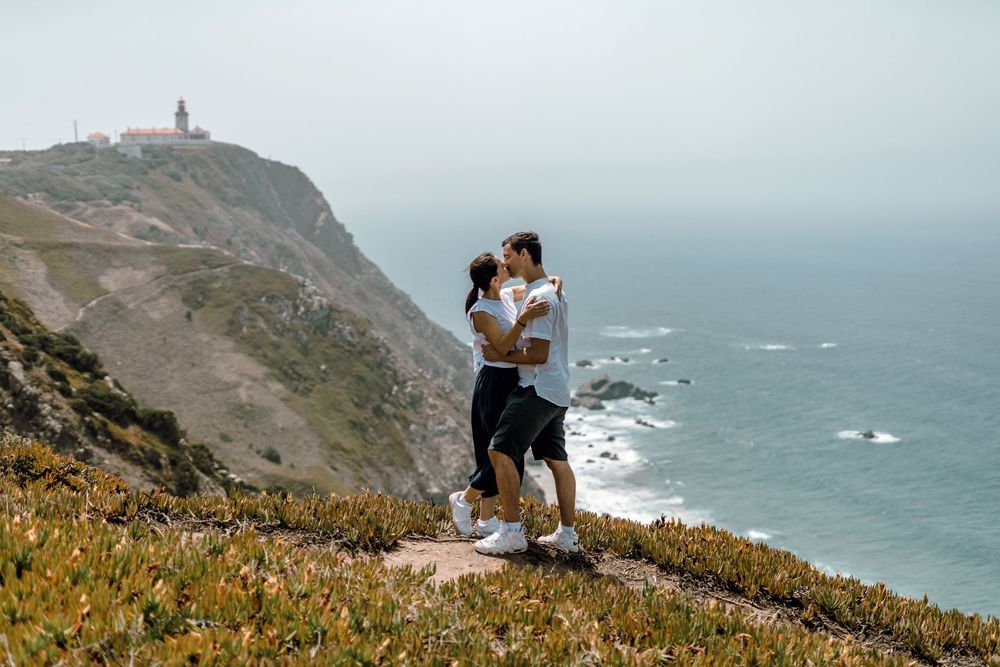 Two people stand on a scenic coastal cliff overlooking the Pacific Ocean with yellow wildflowers in the foreground.