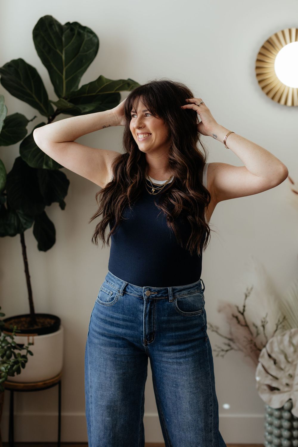 Woman in dark tank top and jeans poses playfully against white wall with indoor plants and decor.