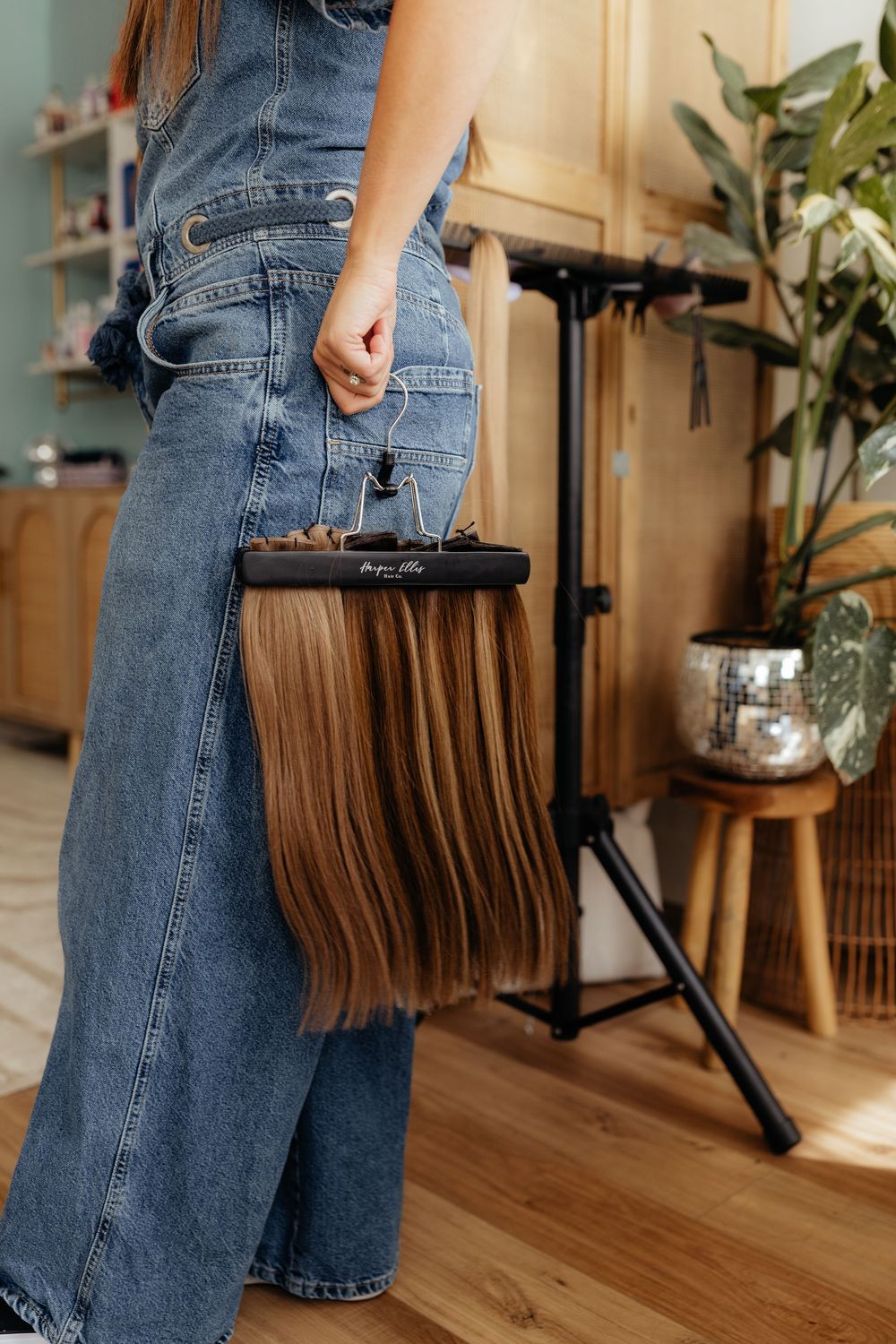 Woman wearing blue jeans holds a brown leather fringed handbag in a sunlit room with wooden floors and plants.