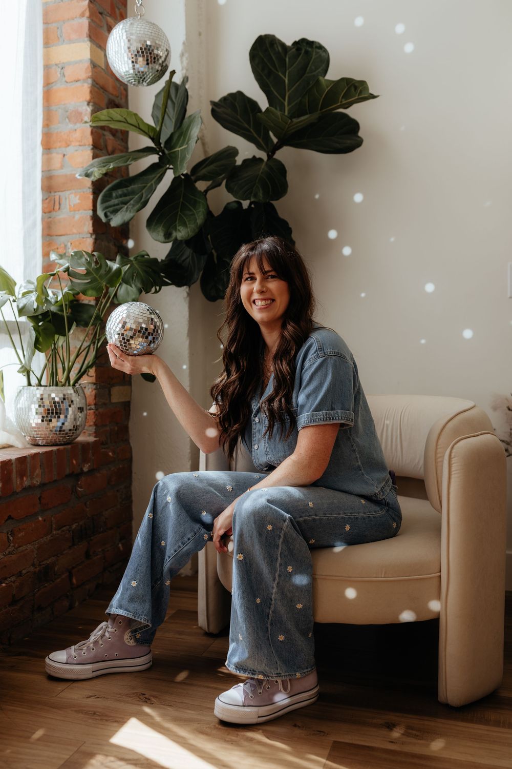 Person in denim sitting on beige armchair with disco ball and fiddle leaf fig plant in modern room interior.
