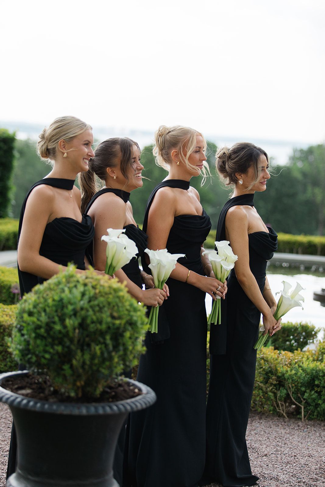 Four bridesmaids in elegant black strapless gowns stand in a row holding white calla lily bouquets in an outdoor garden setting.