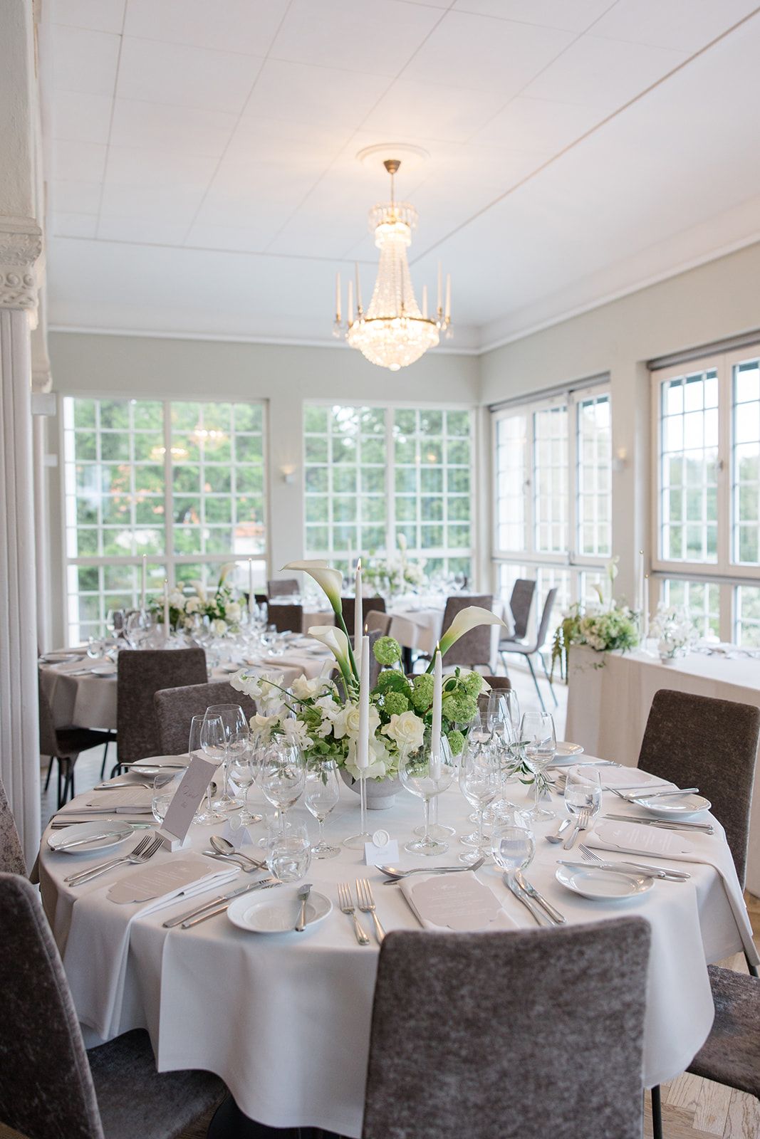 Elegant dining room with white tablecloths, gray chairs, and crystal chandeliers in a bright sunlit space with large windows.
