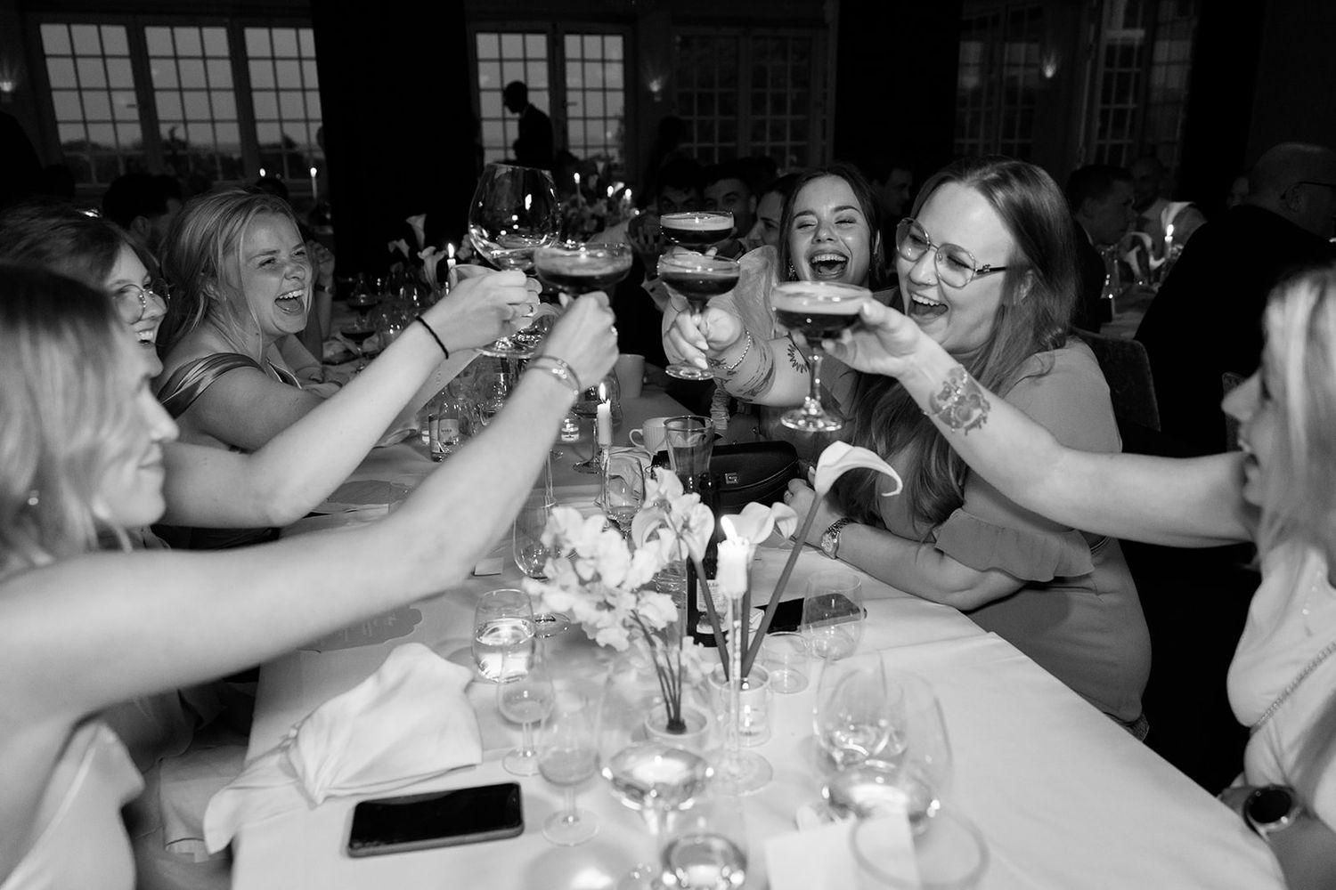 People sitting at tables during a wedding reception share laughter and conversation in a dimly lit venue.