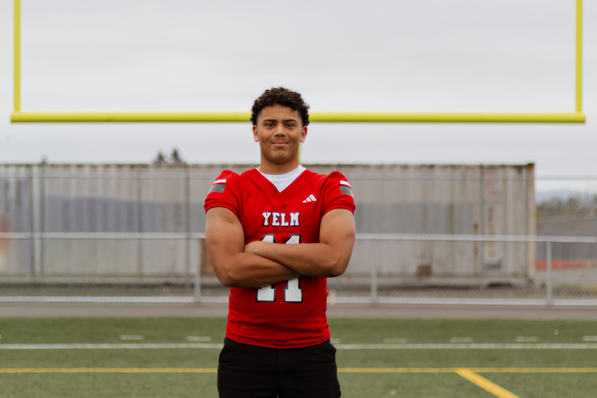 Young male athlete in a red Yelm football jersey number 11 posing with arms crossed on a sports field.