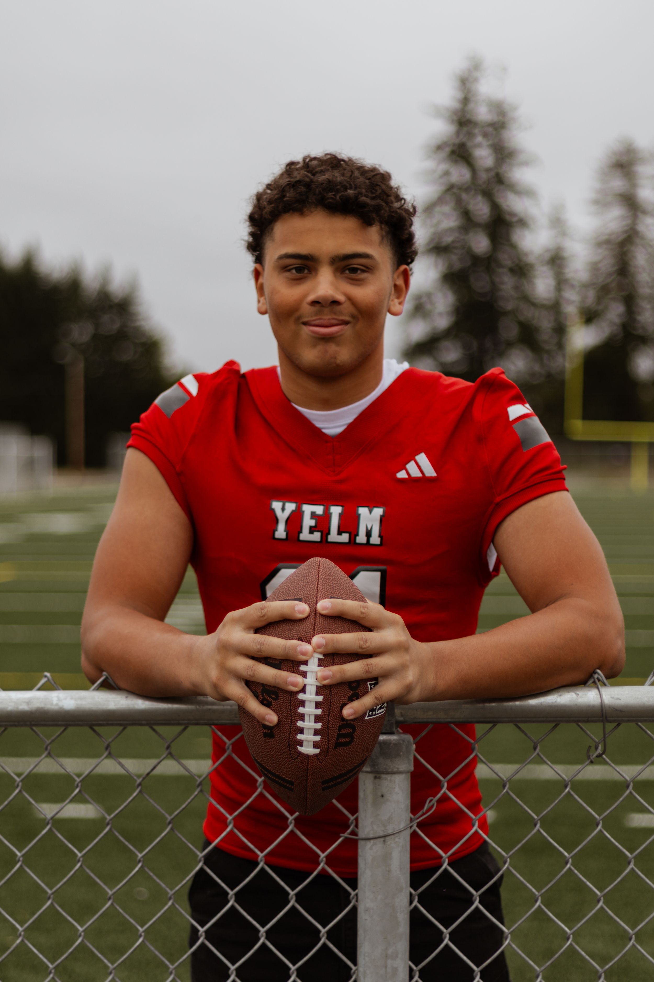 Yelm football player #11 in red jersey holds football while leaning on chain-link fence on field.
