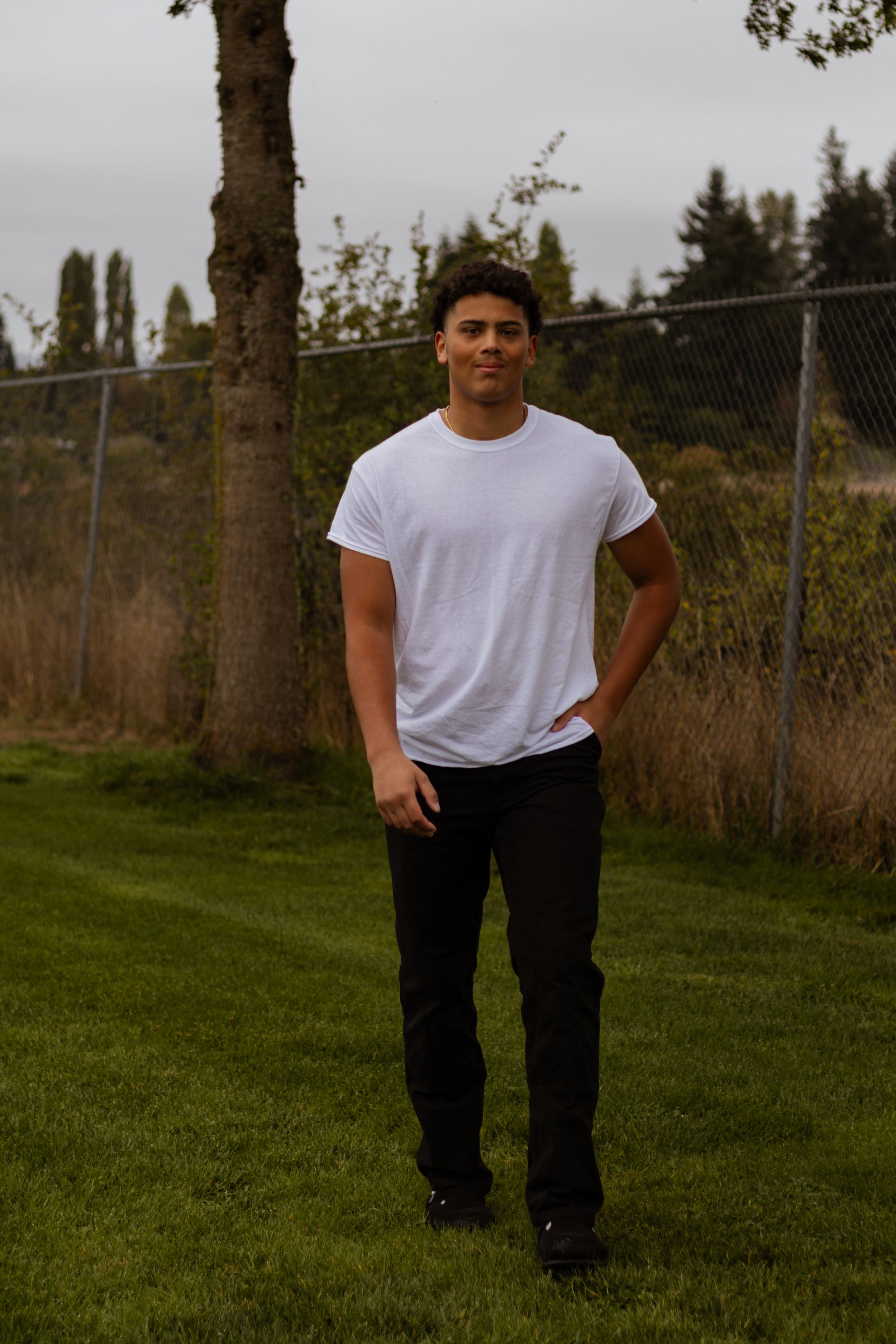 Tall athletic young man in white t-shirt and black pants stands on green grass outdoors.