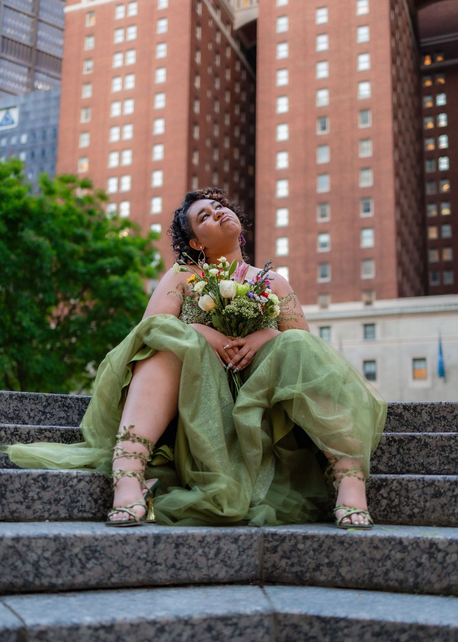 Woman in green formal gown holding bouquet on city steps.