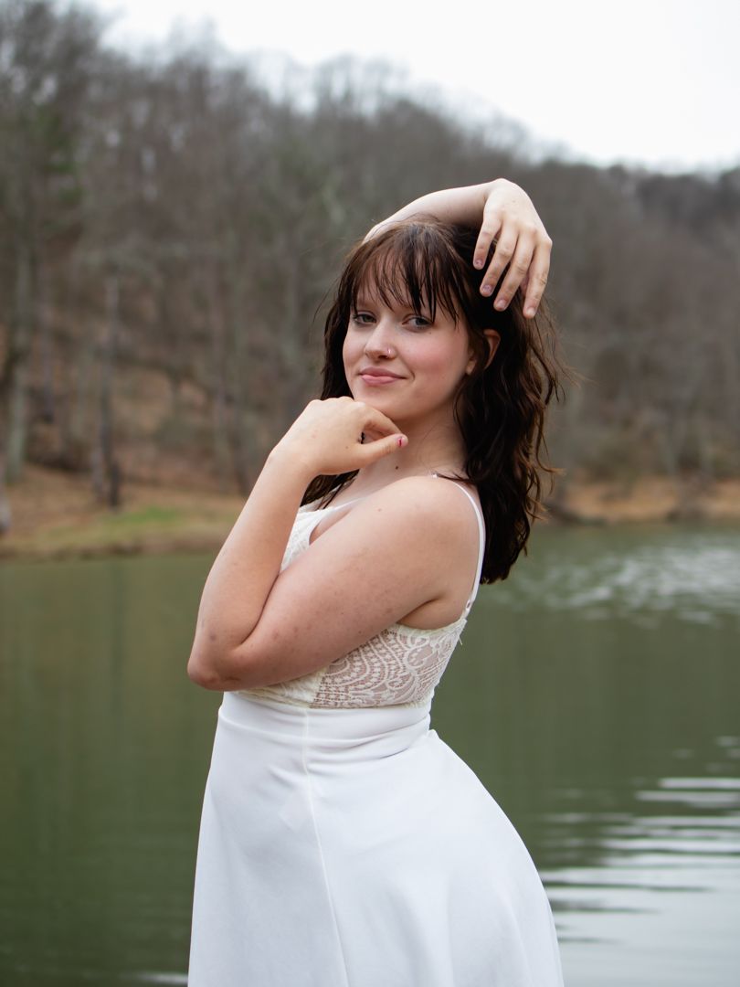 Young woman in white dress posing gracefully by a lake with trees in background.