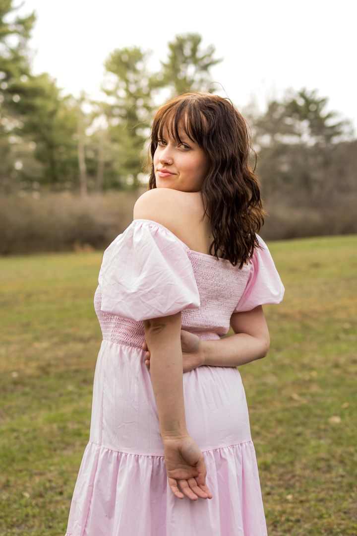 Young woman in pink off-shoulder dress poses outdoors in a green field, looking back over her shoulder.