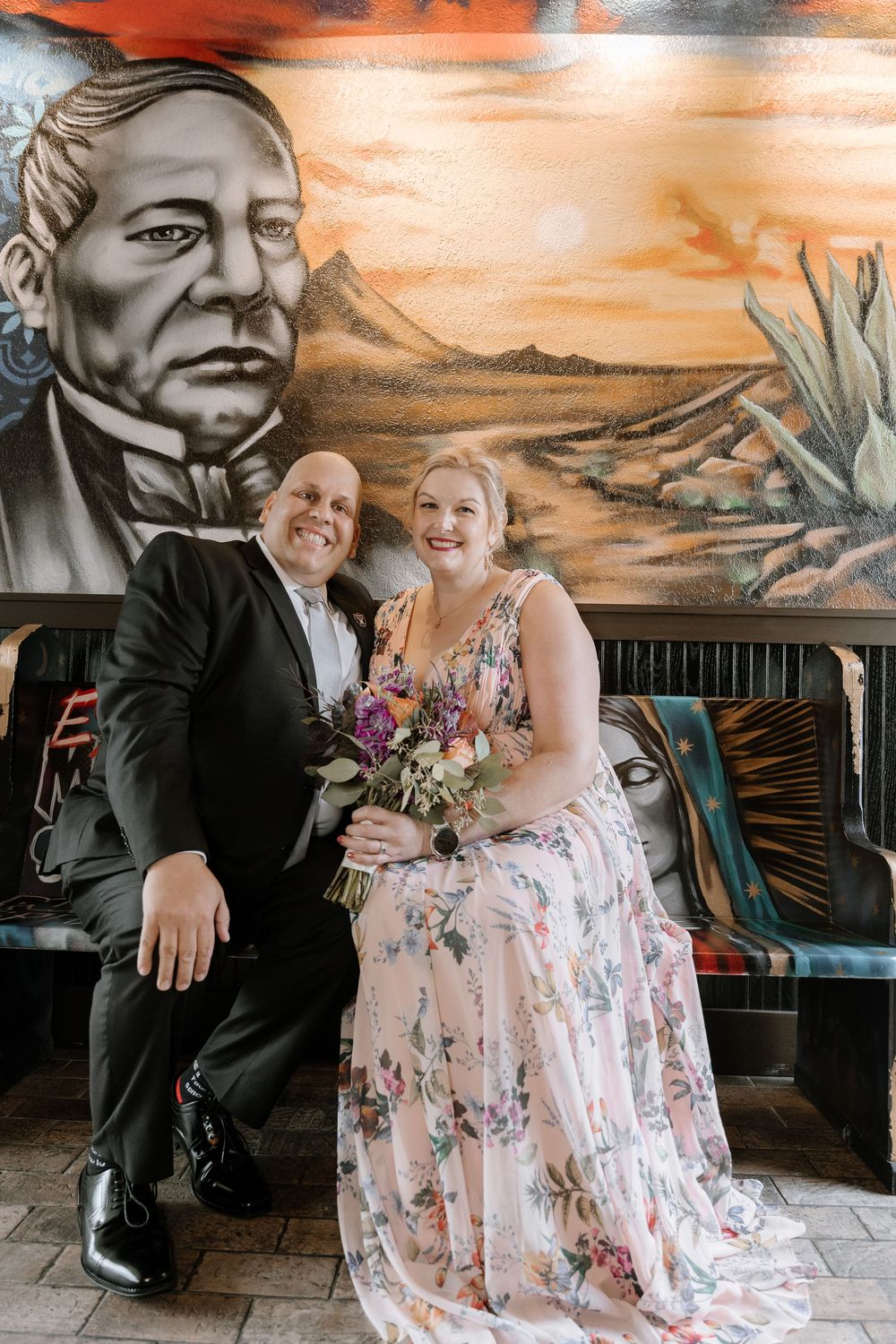 A couple poses against a southwestern-themed mural with desert landscape and cacti in warm orange tones.